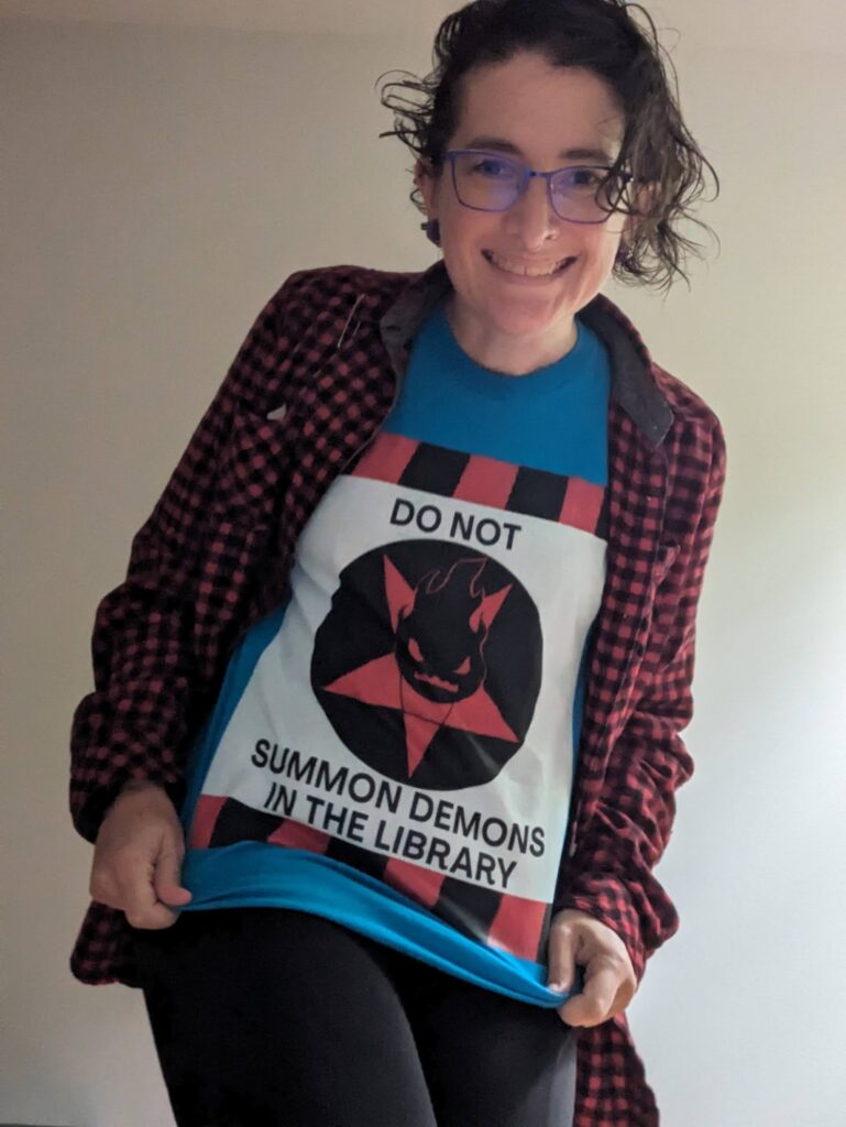 Em wearing a turquoise shirt with a black, red, and white design that says DO NOT SUMMON DEMONS IN THE LIBRARY with a little flame-headed demon.