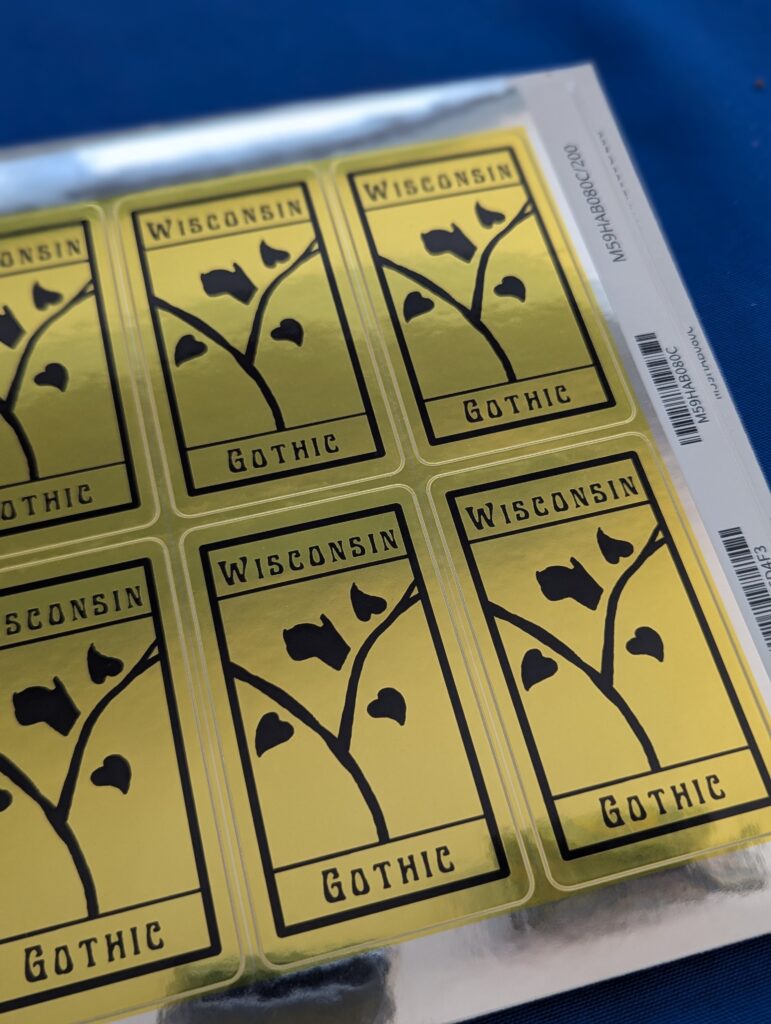 Gold rectangular stickers featuring a vine with three heart-shaped leaves and one Wisconsin-shaped leaf. They say "Wisconsin Gothic."